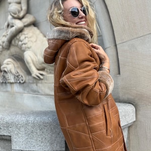 May include: A tan leather coat featuring a patchwork design with visible white stitching, a hood lined with brown faux fur, and matching faux fur trim on the sleeve cuffs. The coat has a long silhouette with side pockets and a smooth finish.