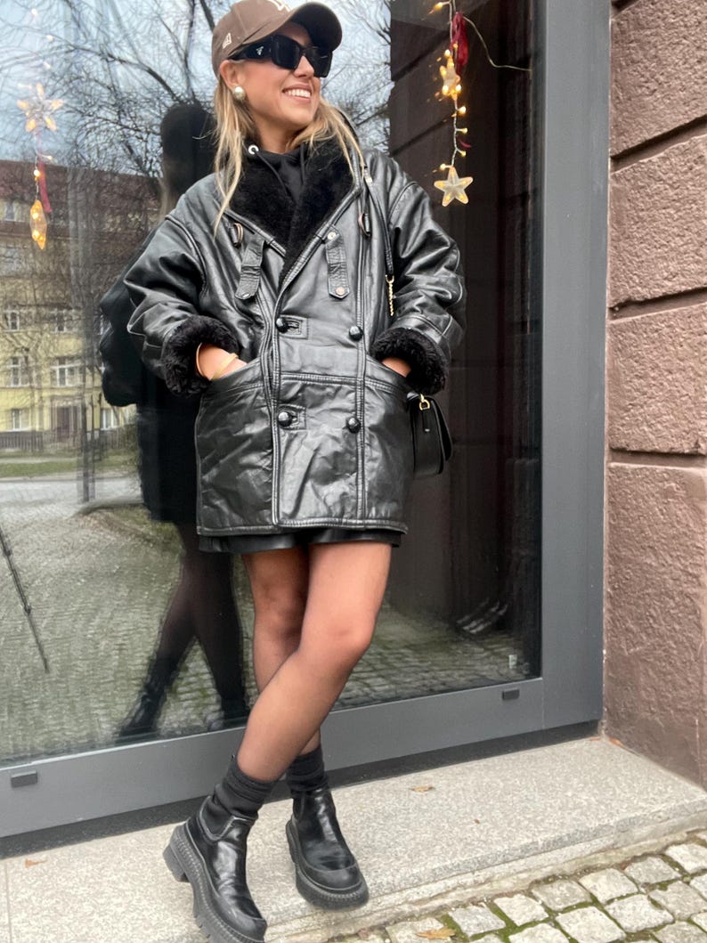 May include: A black leather jacket with a shearling lining and a double-breasted closure. The jacket has a belt at the waist and a hood. The person is wearing black tights and black boots.