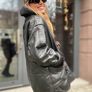 May include: A black leather jacket with a fur-lined collar and cuffs. The jacket has a button closure and a patch pocket on the left side. The person is wearing a brown baseball cap with a white logo and black sunglasses.