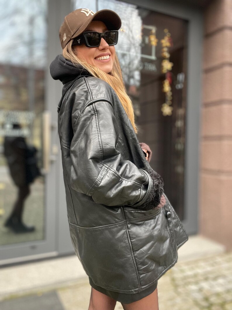 May include: A black leather jacket with a fur-lined collar and cuffs. The jacket has a button closure and a patch pocket on the left side. The person is wearing a brown baseball cap with a white logo and black sunglasses.