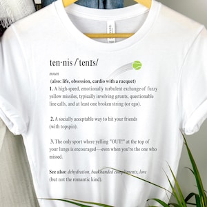 Puede incluir: Camiseta blanca con la definición de tenis impresa en negro. La definición incluye la palabra "sustantivo" y describe el tenis como un intercambio de alta velocidad de misiles amarillos borrosos. También se incluye un gráfico de pelota de tenis verde.