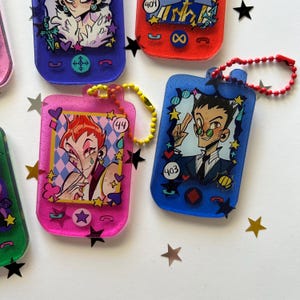 May include: A collection of rectangular resin keychains shaped like vintage mobile phones, featuring colorful glitter finishes and anime-style character illustrations. Each keychain includes a ball chain and displays unique numbers like 44 and 403.