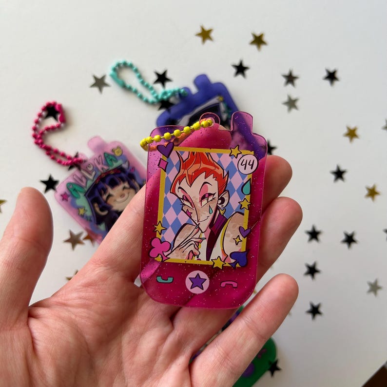 May include: A translucent magenta acrylic keychain shaped like a vintage mobile phone, featuring a printed illustration of a character with spiky orange hair and the number 44. It includes a yellow ball chain and glittery accents throughout.