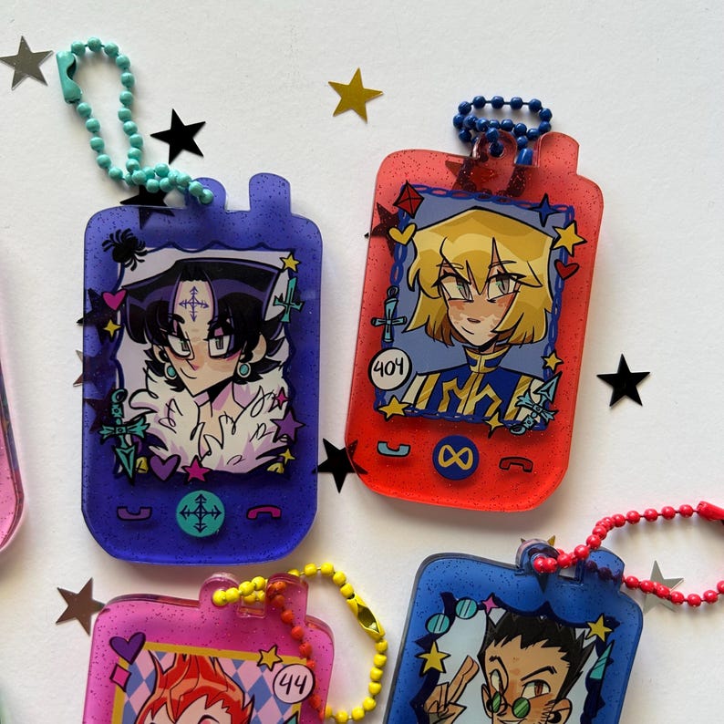 May include: Four rectangular acrylic keychains shaped like vintage handheld devices, each featuring a unique anime-style character illustration. The keychains are translucent with glitter, colored purple, red, pink, and blue, and include matching ball chains.