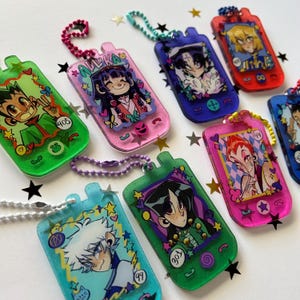 May include: A selection of vibrant, glittery phone-shaped keyrings showcasing anime-style character illustrations. Each keyring is a different colour and character, with details such as stars and numbers. The keyrings are attached to beaded chains.