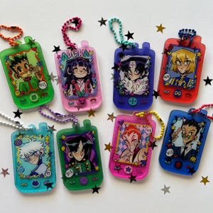 May include: A collection of eight translucent acrylic keychains shaped like vintage handheld game consoles, each featuring a unique anime-style character illustration, colorful ball chains, and small decorative star-shaped cutouts.