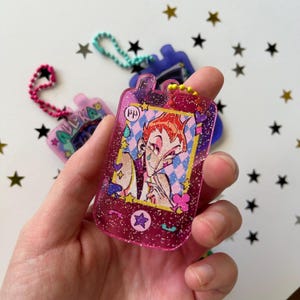 May include: A translucent pink glitter acrylic keychain shaped like a vintage mobile phone. The center features an illustration of a character with orange hair, a teardrop mark, and a star mark on their face, set against a blue and white diamond pattern.