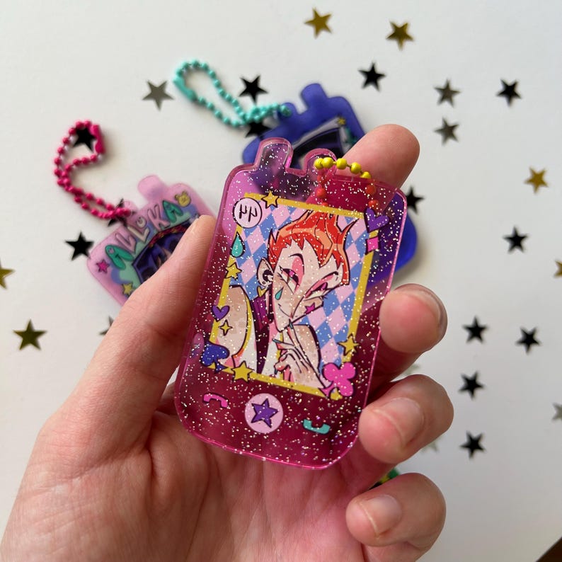 May include: A translucent pink glitter acrylic keychain shaped like a vintage mobile phone. The center features an illustration of a character with orange hair, a teardrop mark, and a star mark on their face, set against a blue and white diamond pattern.
