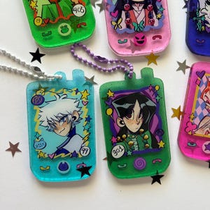 May include: A collection of rectangular resin keychains shaped like handheld digital pets. Each features a colorful glitter finish, a character illustration, and printed numbers like 99, 301, and 405. The keychains include metal ball chains and decorative star-shaped confetti accents.