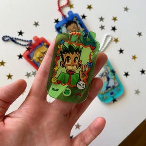 May include: A translucent green acrylic keychain shaped like a vintage mobile phone, featuring a cartoon character illustration, a small frog icon, and the number 405. The keychain includes an orange beaded loop and a glittery finish throughout.