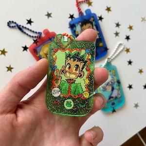 May include: A green glittery resin keychain shaped like a handheld digital pet device, featuring an illustration of a spiky-haired character making a peace sign. The screen area includes small star, heart, and frog icons, with a blue ball chain attached.
