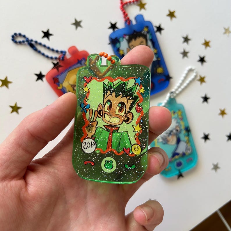 May include: A green glittery resin keychain shaped like a handheld digital pet device, featuring an illustration of a spiky-haired character making a peace sign. The screen area includes small star, heart, and frog icons, with a blue ball chain attached.