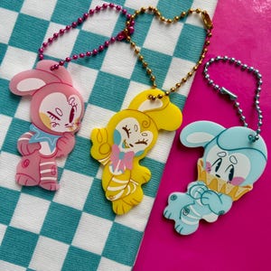 Puede incluir: Tres llaveros de acrílico con personajes de conejos de dibujos animados en rosa, amarillo y azul claro. Cada personaje lleva un cuello con volantes y tiene extremidades a rayas. Los llaveros están unidos a cadenas de bolas metálicas.