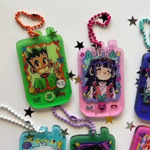 May include: A collection of rectangular resin keychains shaped like vintage mobile phones, featuring glittery finishes and colorful ball chains. Each keychain displays a unique anime-style character illustration, decorative stars, hearts, and small icons.