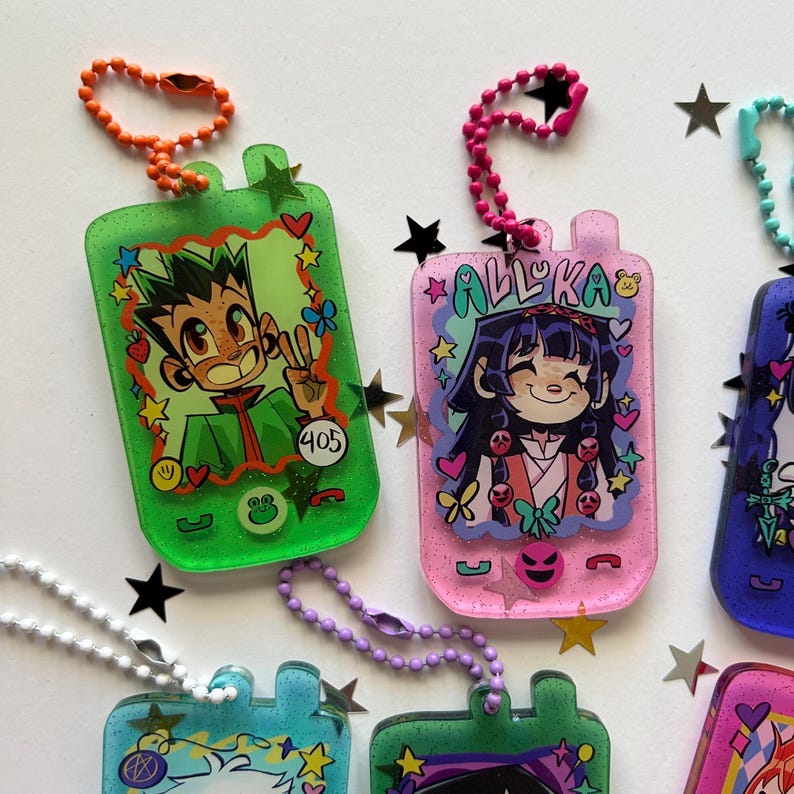 May include: A collection of rectangular resin keychains shaped like vintage mobile phones, featuring glittery finishes and colorful ball chains. Each keychain displays a unique anime-style character illustration, decorative stars, hearts, and small icons.