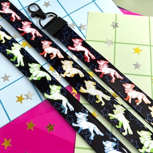Puede incluir: Cordones negros con un patrón repetido de criaturas estilizadas con forma de lobo en varios colores, incluyendo rojo, blanco, amarillo y verde. Cada lobo está sobre un contorno de estrella azul. El cordón tiene un cierre de metal negro.