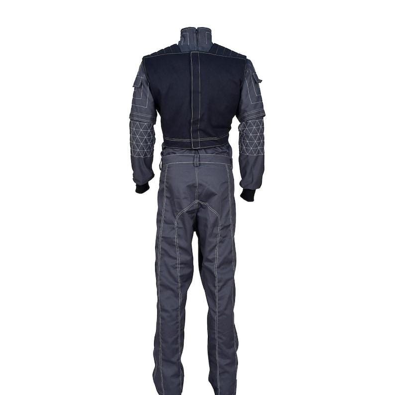 May include: A dark gray and black jumpsuit featuring a high collar, long sleeves with geometric white stitching patterns, and a solid black upper back panel. The garment includes utility-style pockets on the shoulders and white contrast stitching.