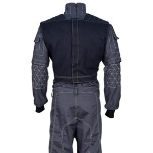 May include: A dark gray and black jumpsuit featuring a high collar, long sleeves with white geometric stitching patterns, and a solid black upper back panel. The garment includes a belt, utility pockets on the shoulders, and white contrast stitching.