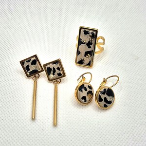 Puede incluir: Un conjunto de joyas doradas con un estampado de manchas en blanco y negro. El conjunto incluye un anillo, dos pares de pendientes, uno con una barra larga y otro con forma redonda. El anillo y los pendientes tienen un marco dorado.