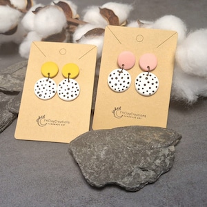 Könnte beinhalten: Zwei Paar handgefertigte Ohrringe aus Polymer Clay. Die Ohrringe sind weiß mit schwarzen Punkten und haben eine gelbe und rosa Oberseite. Die Ohrringe werden auf einer braunen Karte mit dem Text "FKClayCreations handmade art" präsentiert.