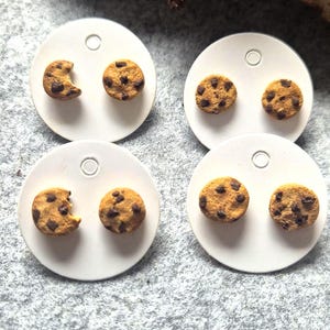 Könnte beinhalten: Vier Paar Ohrstecker in Keksform. Jeder Ohrring ist eine Miniatur-Replik eines Chocolate-Chip-Kekses mit hellbrauner Basis und dunkelbraunen Schokostückchen. Ein Paar hat einen Biss.