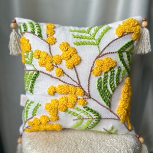 Op de afbeelding: Vierkantig wit decoratief kussen met geborduurde gele mimosa bloemen en groene bladeren. Het kussen heeft bruine stelen, kwastjes en houten kralen op de hoeken. Het kussen ligt op een kleine, witte, gestoffeerde kruk.