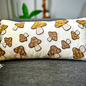 Peut inclure: Un coussin blanc avec des champignons brodés en brun et jaune. Le coussin a des glands à chaque coin.
