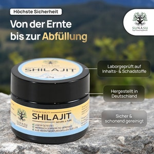 May include: A black jar with a white label that reads "SHILAJIT" in bold, with the words "PREMIUM QUALITY" and "NATURAL & PURE" below. The label also includes a green checkmark next to the words "LABOR GETESTET", "MEHRFACH SORGF&Auml;LTIG GEREINIGT", and "AUS VERANTWORTUNGSVOLLEN QUELLEN". The jar is sitting on a brown rock.