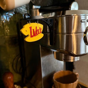 May include: A yellow, coffee-cup-shaped sign with the red text 'Luke's' hanging from a black, ornate, 3D-printed bracket attached to the side of a stainless steel espresso machine. The sign is suspended by two small metal rings from the bracket.