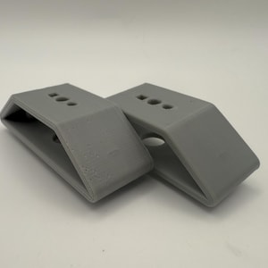 May include: Two gray, angled brackets with a rectangular base and three circular holes on top. The brackets are made of a solid material and have a smooth, matte finish. The brackets are designed to support or connect other objects.