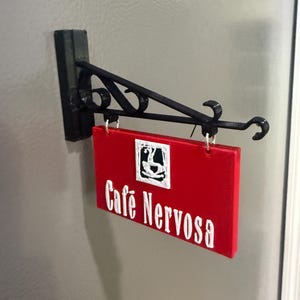 May include: A rectangular red sign with the white text Cafe Nervosa and a white square icon featuring a black coffee cup graphic. The sign hangs from a black metal bracket with decorative scrollwork, mounted to a flat vertical surface.