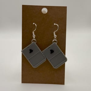May include: A pair of gray earrings with a silver hook. Each earring features a gray, rectangular design with a black heart-shaped accent. The earrings are displayed on a brown card.