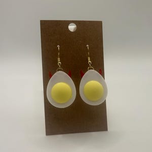 May include: A pair of novelty earrings shaped like fried eggs, each with a yellow yolk and white egg white. The earrings are suspended from gold-colored hooks and have small red devil horns attached.