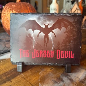 May include: A rectangular slate sign featuring a red illustration of the Jersey Devil with outstretched wings, set against a stormy sky with lightning. The words "The Jersey Devil" are in red. The sign is supported by a metal stand.