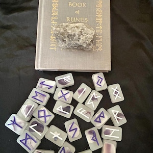 Puede incluir: Un conjunto de piedras rúnicas con símbolos blancos y detalles morados, dispuestas frente a un libro titulado "The Book of Runes". Una piedra gris descansa sobre el libro. El conjunto está sobre una superficie negra.