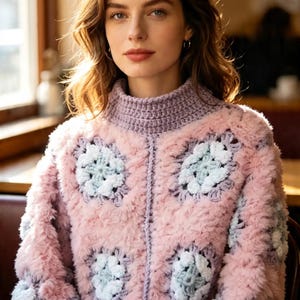 Könnte beinhalten: Ein langärmeliger Pullover mit flauschiger rosa Textur und integrierten hellblauen sowie weißen Häkelmotiven im Granny-Square-Stil. Das Kleidungsstück verfügt über einen hohen Stehkragen und gerippte Bündchen aus einfarbig hellviolettem Garn mit sichtbaren Nähten.