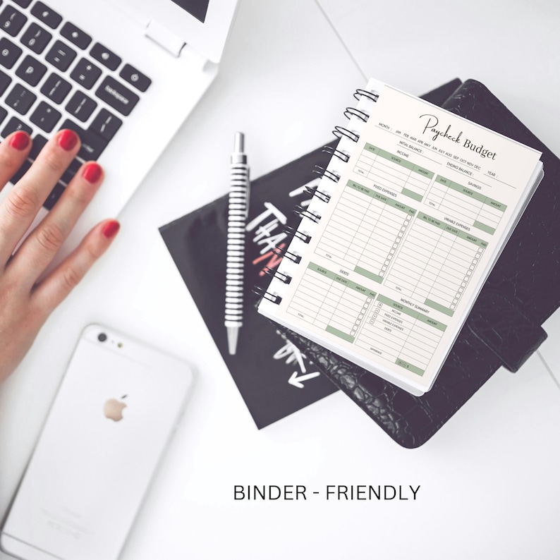 May include: A white spiral bound notebook with the title "Paycheck Budget" on the cover. The notebook is open to a page with a budget tracker. The notebook is on a black surface with a white laptop and a white smartphone in the background.
