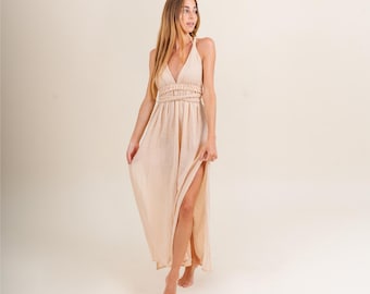 Op de afbeelding: Een licht perzik halter maxi-jurk met een diepe V-hals en een hoge split. De jurk heeft een geplooid taille en een zwierige rok. Het model is blootsvoets. De achtergrond is effen lichtbeige.