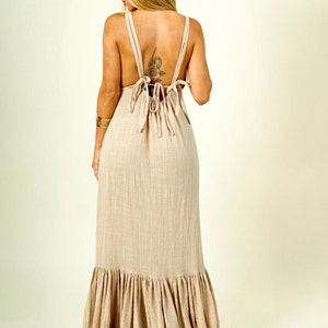 May include: A long, flowy beige maxi dress with a tiered ruffle hem. The dress has a low back with a tie detail.