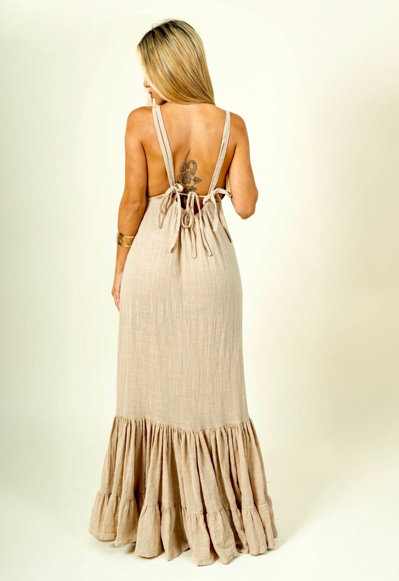 May include: A long, flowy beige maxi dress with a tiered ruffle hem. The dress has a low back with a tie detail.