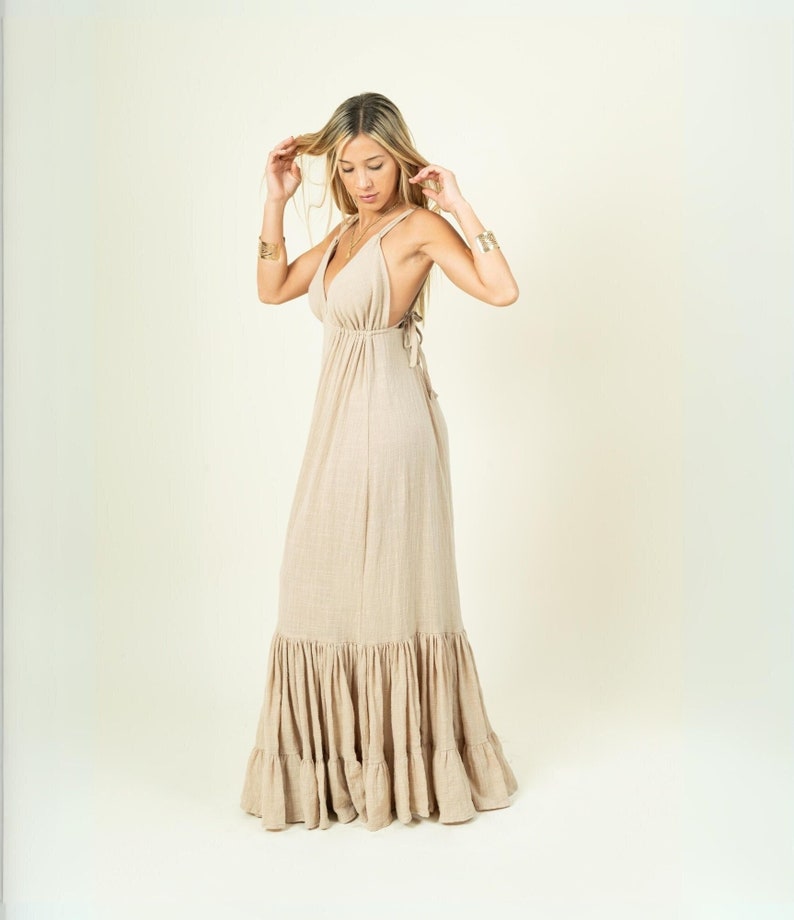 May include: A woman wearing a long, beige maxi dress with a ruffled hem. The dress has a V-neckline and thin straps. The woman is standing on a white background.