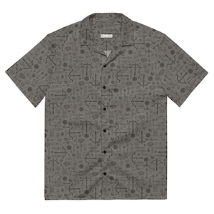 Puede incluir: Camisa gris con botones, cuello de campamento y mangas cortas. La camisa presenta un patrón repetido de ilustraciones negras, que incluyen arcos y flechas, llaves y otros símbolos místicos. La camisa tiene una etiqueta en el cuello.