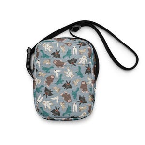 Puede incluir: Bolso bandolera con correa negra y fondo azul claro. El bolso está cubierto con un estampado de varios críptidos de dibujos animados, incluyendo Bigfoot, murciélagos y fantasmas. El bolso tiene una hebilla negra.