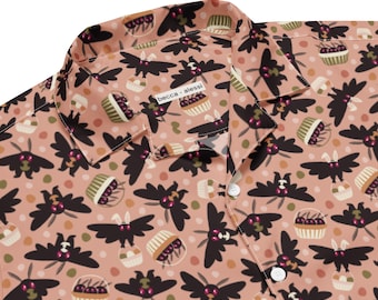 May include: A button-down shirt with a repeating pattern of black bats and cupcakes on a peach background. The shirt has a collar and white buttons. The brand name "becca alessi" is printed on the collar.
