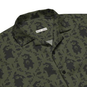 May include: An olive green button-down shirt featuring a repeating dark gray pattern of plague doctor figures, small rats, and medical syringes. The collar displays a white rectangular label with the black text becca • alessi printed on it.