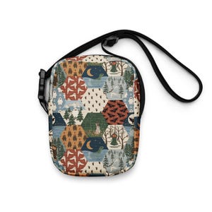 May include: A crossbody bag featuring a repeating hexagonal pattern with illustrations of cryptids, forest scenes, and nocturnal elements in muted earth tones. The bag includes a black adjustable strap with a plastic buckle closure mechanism.