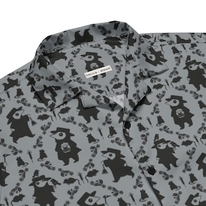 May include: A gray button-down shirt featuring a repeating black pattern of plague doctor figures, rats, syringes, and small birds. The collar displays a white rectangular label with the black text becca alessi printed on the fabric.