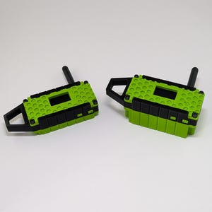 Op de afbeelding: Twee rechthoekige elektronische apparaten in fel limoengroen en zwart. Elke eenheid heeft een gestructureerd honingraatpatroon op het bovenoppervlak, een zwarte antenne, een handvat aan de zijkant en een kleine rechthoekige uitsparing voor een scherm.