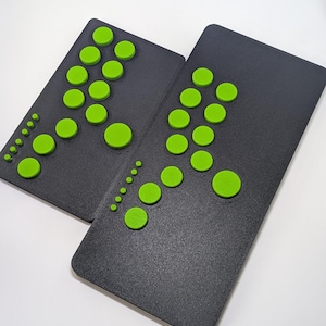 Può includere: Due controller di gioco rettangolari grigio scuro con una superficie testurizzata e molteplici pulsanti circolari verde lime brillante di varie dimensioni disposti secondo uno schema specifico sulla faccia di ogni dispositivo.