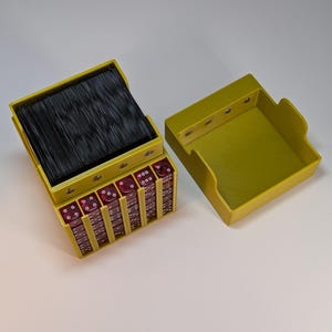 Könnte beinhalten: Eine gelbe Plastikbox mit einem schwarzen Kartenspiel und roten Würfeln im Inneren. Die Box ist geöffnet und die Würfel sind sichtbar. Die Box ist so konzipiert, dass sie ein Kartenspiel und Würfel aufnehmen kann.